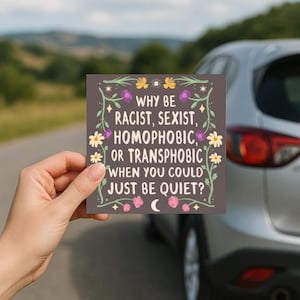 May include: A square sticker with the text "WHY BE RACIST, SEXIST, HOMOPHOBIC, OR TRANSPHOBIC WHEN YOU COULD JUST BE QUIET?" is held up in front of a car. The sticker has a floral border with yellow, purple, and pink accents.