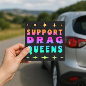 May include: A square sticker with the text "SUPPORT DRAG QUEENS" in colorful, gradient lettering. The sticker is held in front of a car, with a blurred background of trees and a road.