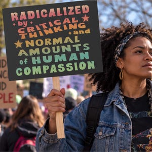 以下が含まれることがあります： ダークグレーの背景に多色のテキストで「RADICALIZED by CRITICAL THINKING and NORMAL AMOUNTS of HUMAN COMPASSION」と書かれた長方形の抗議用サイン。サインは、人が持っている明るい茶色の木製の持ち手に取り付けられています。