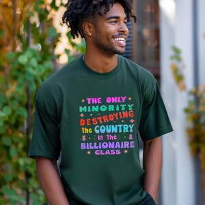 Könnte beinhalten: Dunkelgrünes T-Shirt mit dem Text "THE ONLY MINORITY DESTROYING THE COUNTRY is the BILLIONAIRE CLASS" in bunten, stilisierten Buchstaben. Das T-Shirt wird von einer lächelnden Person getragen.