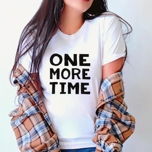 Peut inclure: T-shirt blanc avec le texte "ONE MORE TIME" en lettres noires en gras. La personne porte une chemise à carreaux marron et bleue par-dessus le t-shirt.