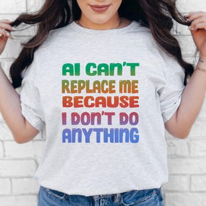 May include: A light gray short-sleeved t-shirt featuring a multi-colored gradient graphic print with the text AI CAN'T REPLACE ME BECAUSE I DON'T DO ANYTHING. The lettering transitions from green to orange, red, and blue in a blocky, distressed font.