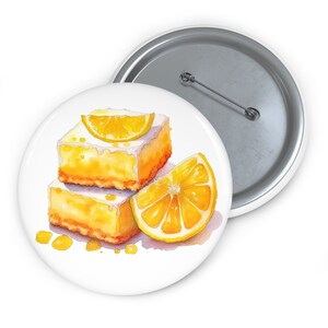 May include: A round pin-back button featuring a watercolor illustration of two stacked lemon bars topped with a fresh lemon slice, accompanied by an additional lemon wedge and small yellow droplets on a white background with a metal pin back.