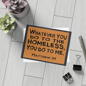 May include: A greeting card with the text "WHATEVER YOU DO TO THE HOMELESS, YOU DO TO ME. MATTHEW 25" in black on a brown background. The card is on a white surface with a black border. The image also includes two small potted succulents, a pen, and binder clips.