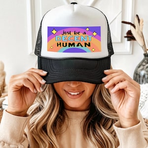 Op de afbeelding: Zwarte en witte truckerpet met een mesh achterkant en een wit voorpaneel. Het voorpaneel heeft een kleurrijke afbeelding met de tekst "Just be a DECENT HUMAN" boven een regenboogontwerp.