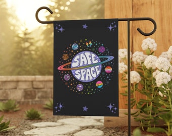May include: A black garden flag with a rainbow colored planet and the text "SAFE SPACE" in a retro font. The planet is surrounded by smaller planets and stars.