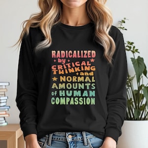 Puede incluir: Camiseta negra de manga larga con el texto "RADICALIZED by CRITICAL THINKING and NORMAL AMOUNTS of HUMAN COMPASSION" en letras coloridas de estilo retro. Una prenda cómoda e informal.