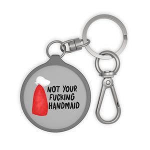 May include: A round, gray keychain with a silver clasp and ring. The keychain features the text "NOT YOUR FUCKING HANDMAID" in black, along with a red and white graphic. The keychain is a novelty item.