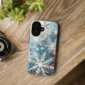 Puede incluir: Una funda para teléfono con un diseño de acuarela de copos de nieve sobre un fondo azul. La funda tiene un recorte para la cámara y un borde blanco.
