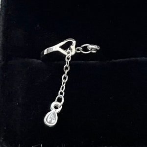 May include: A silver ring with a heart-shaped design and a dangling chain with a small clear stone at the end. The ring is in a black velvet ring box.