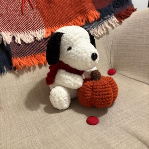 May include: A handmade crochet Snoopy doll with black ears, a red scarf, and a small orange pumpkin. The plush toy is sitting on a beige chair with a colorful blanket in the background. Red circular embellishments are scattered around.
