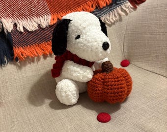 May include: A handmade crochet Snoopy doll with black ears, a red scarf, and a small orange pumpkin. The plush toy is sitting on a beige chair with a colorful blanket in the background. Red circular embellishments are scattered around.