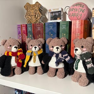 May include: Four handmade crochet teddy bears wearing black robes and scarves in different colors. The bears are arranged on a white shelf in front of a collection of books and decorative items. The text "HAPPEE BIRTH DAE HARRY" is visible.