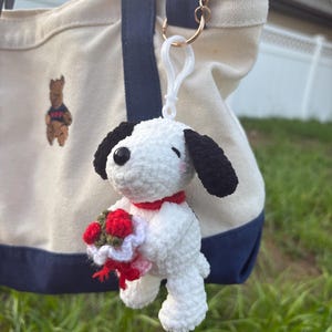 May include: A white crocheted dog keychain with black ears and a red collar, holding a small bouquet of red flowers with green leaves. The figure is attached to a white plastic clip and a gold metal ring, hanging from a cream-colored canvas tote bag.