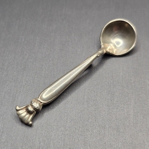 May include: A small, silver spoon with a rounded bowl and a decorative handle. The handle features a detailed design at the base and tapers towards the bowl. The spoon is likely made of metal and is displayed against a gray background.