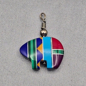 May include: A bear-shaped pendant featuring inlaid segments of blue, green, yellow, turquoise, red, and purple stone. The pendant is accented with thin silver-colored metal strips between the stones and includes a silver-colored metal bail at the top.