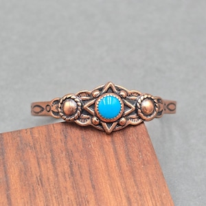 May include: A copper-toned bracelet with a central turquoise-colored stone. The bracelet features an ornate design with a star-shaped centerpiece and circular accents. The bracelet is resting on a wooden surface.