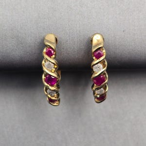 May include: A pair of gold hoop earrings featuring a twisted design. Each earring is set with alternating round-cut vibrant red rubies and clear white diamonds, creating a repeating pattern along the curved surface of the metal band.