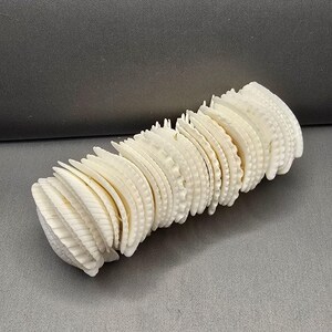 May include: A cylindrical decorative object constructed from a series of stacked, off-white, scalloped-edge seashell discs. The individual shells are arranged in a tight, repeating pattern to form a solid, elongated, tube-like structure.