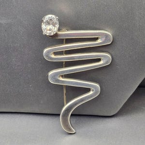 Op de afbeelding: Een zilverkleurige metalen broche met een doorlopende, golvende zigzagvorm die naar onderen toe smaller wordt. Een enkele, ronde, heldere gefacetteerde steen is in de linkerbovenhoek van de metalen structuur gezet met klauwtjes.