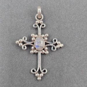 Peut inclure: Un pendentif croix en argent orné d'une pierre de lune ovale en son centre. La structure métallique présente des volutes décoratives, de petites perles sphériques et une bélière circulaire au sommet pour l'attacher à une chaîne.
