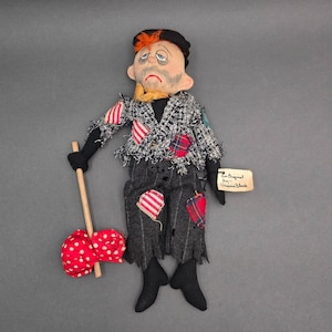 Puede incluir: Una muñeca de tela con cara pintada, pelo naranja y gorra negra. Viste una chaqueta gris deshilachada con parches de rayas rojas y blancas, pantalones gris oscuro con agujeros y lleva un palo de madera con un hatillo de lunares rojos y blancos.