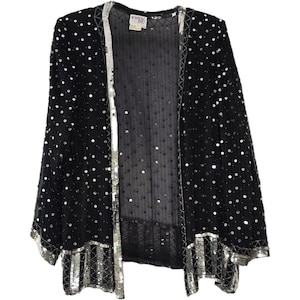 Könnte beinhalten: Eine schwarze, vorne offene Jacke aus transparentem Netzstoff, verziert mit verstreuten silbernen Pailletten. Das Kleidungsstück hat einen breiten, dicht mit silbernen Pailletten bestickten Rand an der Öffnung, den Ärmelbündchen und am Saum, mit Etikett ROUGE ET NOIR.