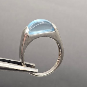 May include: A silver ring with a large, light blue, oval-shaped stone. The ring has a simple, modern design with a smooth, polished finish. The band is thick and sturdy, with small details on the side.