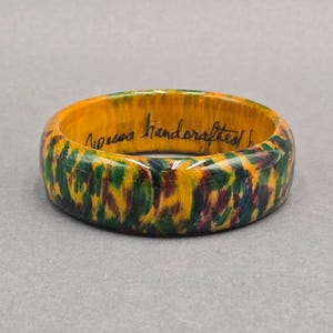 May include: A circular bangle bracelet featuring a vibrant, multi-colored pattern of yellow, green, and dark brown brushstrokes. The interior of the bracelet is a solid yellow color with the black handwritten text "cypress handcrafted" visible.
