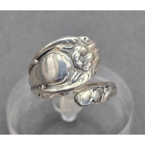 May include: A silver-toned metal ring crafted from a repurposed spoon handle. The ring features an open-ended bypass design with an embossed floral motif on the wider end and intricate scrollwork detailing along the curved band surface.