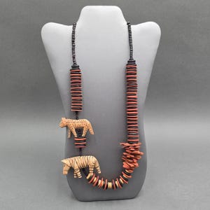 May include: A handcrafted necklace featuring a series of stacked, round, coral and black beads. Two carved animal pendants, a leopard and a tiger, are suspended from the necklace. The necklace is displayed on a gray mannequin.
