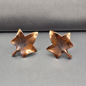 May include: A pair of copper-colored leaf-shaped earrings. Each earring has a detailed leaf vein design and a small stem. The earrings are displayed on a gray surface.