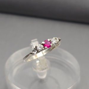 May include: A silver ring featuring a central, round, bright red gemstone flanked by two clear, round gemstones on each side. The ring is set on a clear display stand against a gray background. The ring's band is thin and appears to be made of silver.