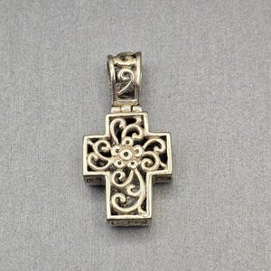 Op de afbeelding: Zilverkleurige kruisvormige hanger met een sierlijk, opengewerkt ontwerp. Het kruis heeft een bloemenmiddelpunt en krullende details. De beugel is ook versierd met een bijpassend patroon. Een religieus sieraad.