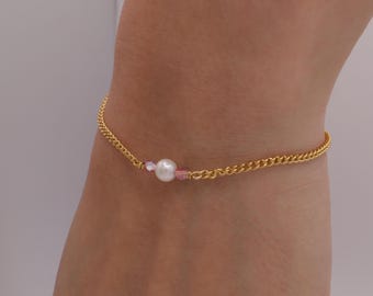 May include: A gold-colored chain anklet with a central white pearl and two pink beads. The delicate jewelry piece is designed to be worn around the ankle. The chain has a curb link design.