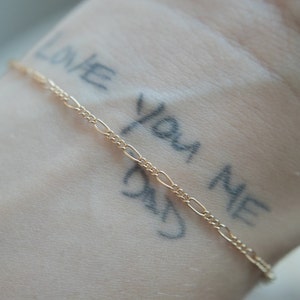 Peut inclure: Un bracelet en chaîne dorée sur le poignet d'une personne avec les mots "Love You Me Day" écrits sur sa peau.