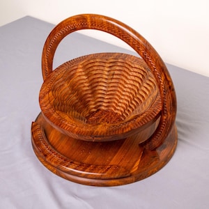 May include: A wooden fruit basket with a carved, wavy interior and a curved handle. The basket is made of a warm, reddish-brown wood and sits on a round base. The handle is also carved with a decorative pattern.