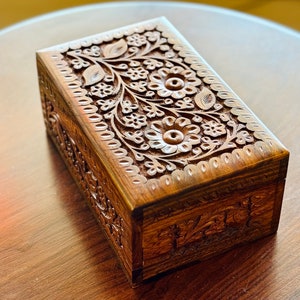 May include: A wooden box with intricate floral carvings. The box is made of dark wood and has a hinged lid. The carvings are detailed and cover the entire surface of the box.