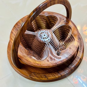 May include: A handcrafted wooden serving basket with a handle. The basket features a tiered design with carved floral and leaf patterns. The wood has a warm, brown tone, and the handle arches gracefully above the basket.