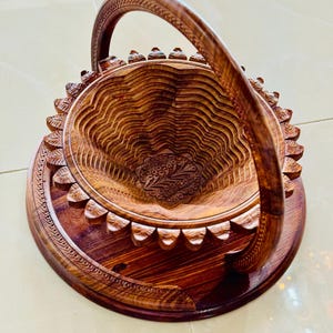 May include: A handcrafted wooden basket with a carved floral design. The basket features a round base, a scalloped edge, and a curved handle. The wood has a rich, warm brown color, and the intricate carvings add a touch of elegance.