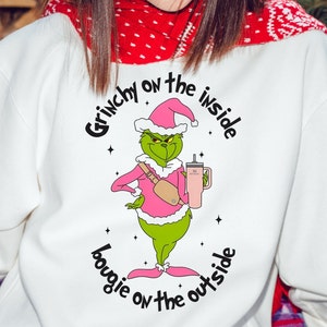 Puede incluir: Sudadera blanca con un personaje verde del Grinch que lleva un traje de Papá Noel rosa y sostiene una taza rosa. El texto en la sudadera dice "Grinchy on the inside, bougie on the outside."