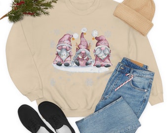 Op de afbeelding: Een beige sweatshirt met een afbeelding van drie gnomen die roze mutsen dragen en lichtjes vasthouden. De gnomen staan op een besneeuwde achtergrond met sneeuwvlokken.