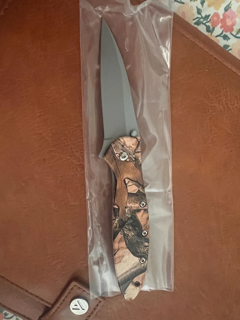May include: A folding knife with a grey blade and a camouflage handle. The knife is in a clear plastic sleeve. The handle features a brown, tan, and black camouflage pattern. The knife is designed for outdoor use.