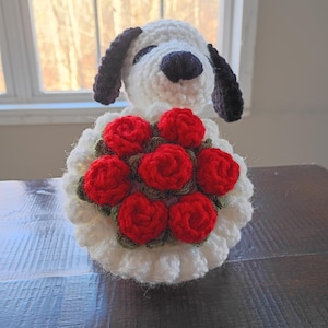 May include: A handmade crochet dog with a white body, black ears, and a black nose. The dog is holding a bouquet of red roses with green leaves. The dog and roses are on a dark wooden surface.