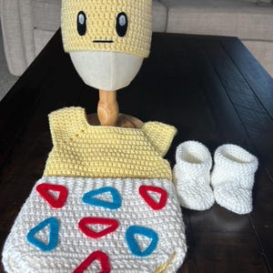 K&ouml;nnte beinhalten: Handgefertigtes Baby-Set aus H&auml;kelarbeit in Gelb und Wei&szlig;, bestehend aus Hut, Strampler und Babyschuhen. Der Strampler hat rote und blaue Dreiecksakzente. Der Hut ist mit einem Cartoon-Gesicht gestaltet.