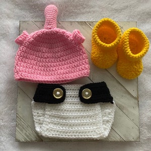 May include: A crocheted baby outfit set in pink, white, and black with yellow booties. The set includes a hat, diaper cover, and booties.