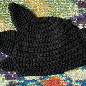 May include: Black crocheted hat with a pointed top and ear flaps. The hat is made with a textured stitch.
