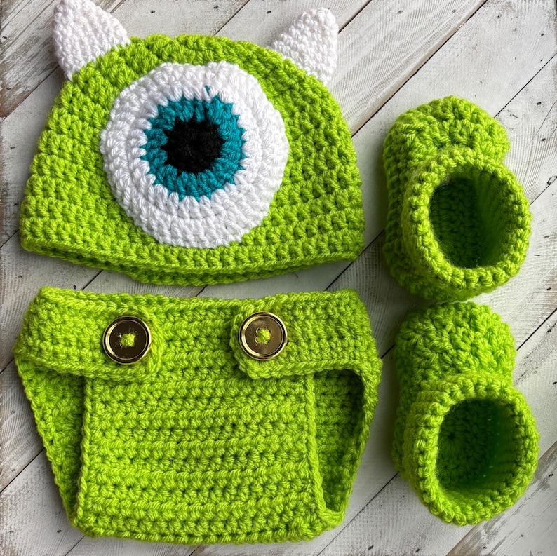 May include: A lime green crochet baby set featuring a hat with a large eye design, a diaper cover with gold buttons, and two booties. The hat has white ears and a blue, white, and black eye. The set is handmade.