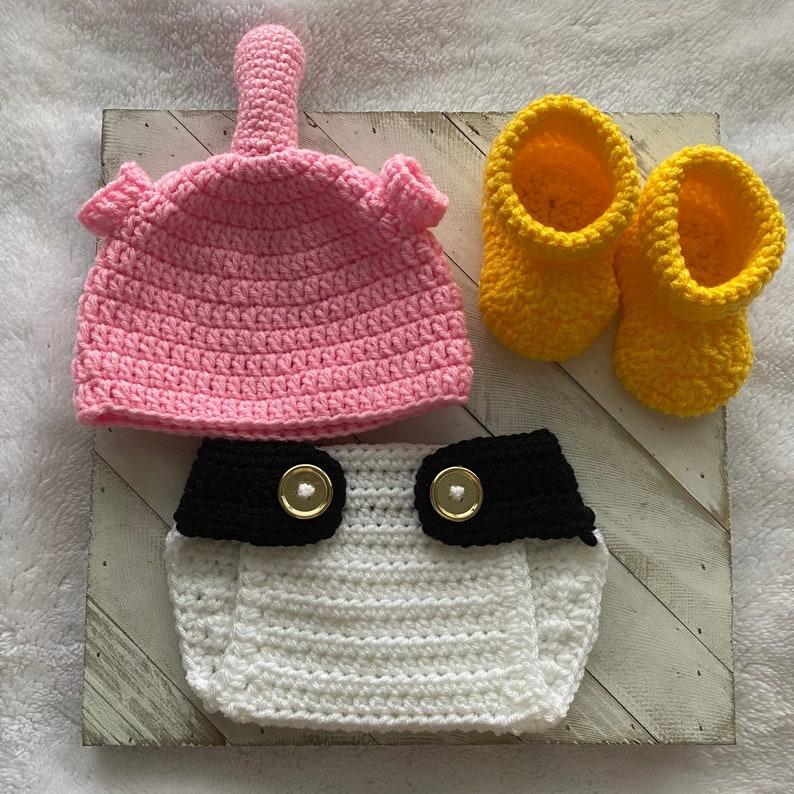 May include: A crocheted baby outfit set in pink, white, and black with yellow booties. The set includes a hat, diaper cover, and booties.