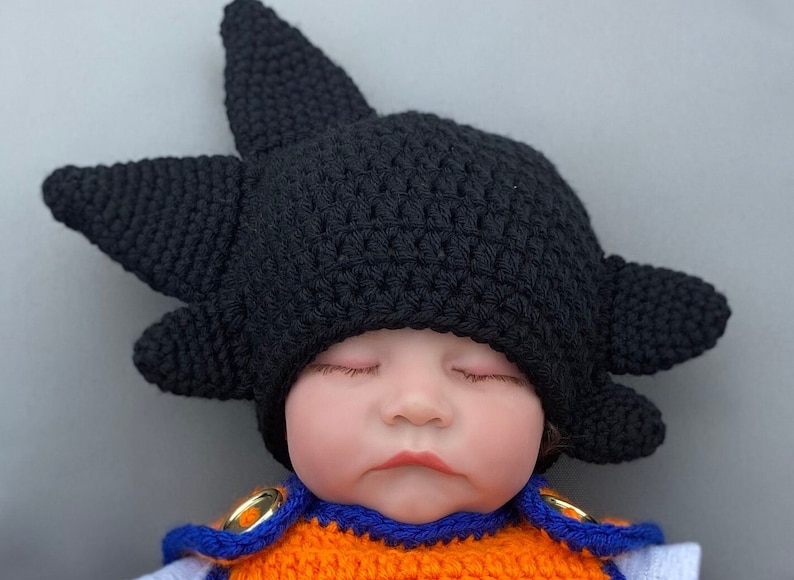 May include: A sleeping baby wearing a black crocheted hat with spiky points, resembling a popular anime character's hair. The baby is also wearing an orange and blue crocheted outfit.